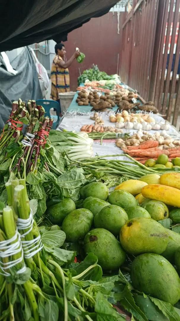 A market stall laid out with greens, mangoes, root crops and vegetables